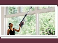 A woman using a cordless vacuum to clean her window sills. She's reaching a sill above her head.
