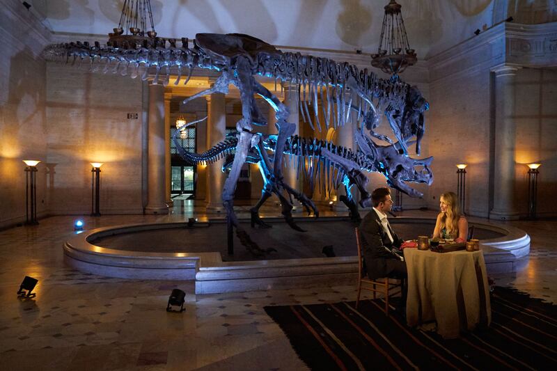 Zach and Kaity sit in a museum in front of a skeleton of a dinosaur