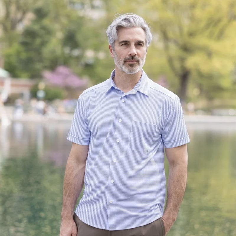 Man wearing a light blue striped short-sleeve button-down shirt, standing in front of a reflective pond in a park setting.