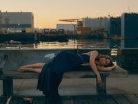 A woman lies on a bench in a blue dress near the water.