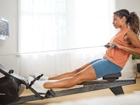 A woman uses a Hydrow Wave rowing machine in a bright home gym, pulling the handle as she follows an instructor on the built-in screen.