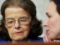 A picture of U.S. Senator Dianne Feinstein (D-CA) listening to an aide. She was recently reminded to “just say aye” when asked to vote on the defense appropriations bill.