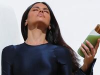 A model in a navy dress holds a glass bottle of green gut powder drink mix with a gold cap, posed against a white background.