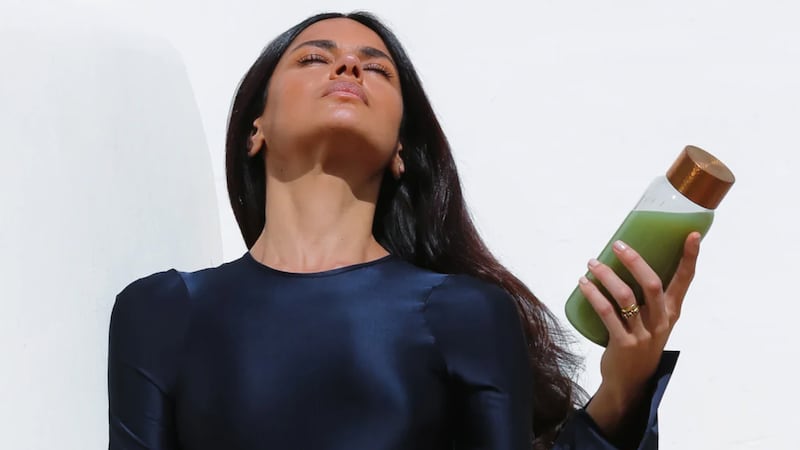 A model in a navy dress holds a glass bottle of green gut powder drink mix with a gold cap, posed against a white background.
