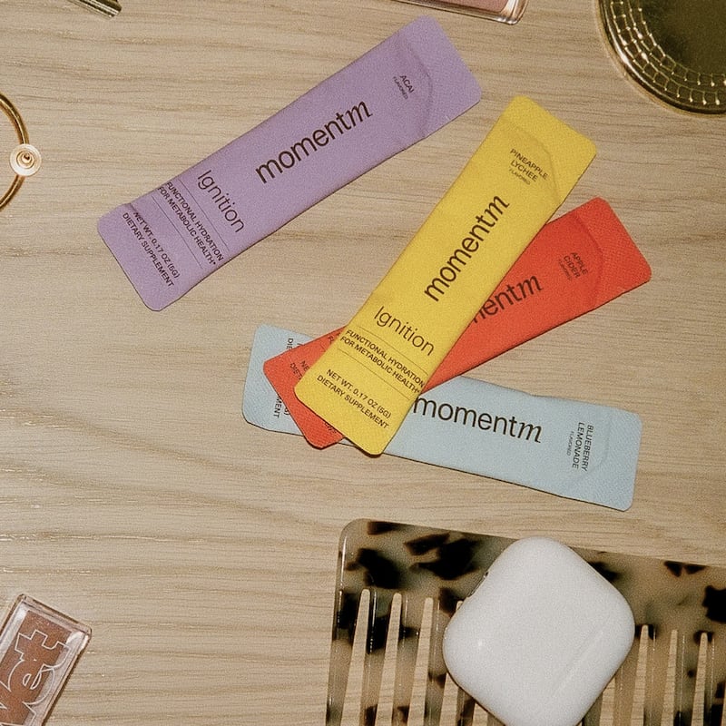 Four momentm Ignition hydration packets in açai, pineapple lychee, apple cider, and blueberry lemonade flavors, arranged on a wooden surface alongside gold jewelry, a tortoiseshell comb, and wireless earbuds.