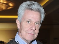 A grey-haired man holds an action figure of a man in a gimp costume.