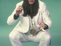 a vintage photo of rock star Ozzy Osbourne in a white suit with long straight black hair and black eyeshadow