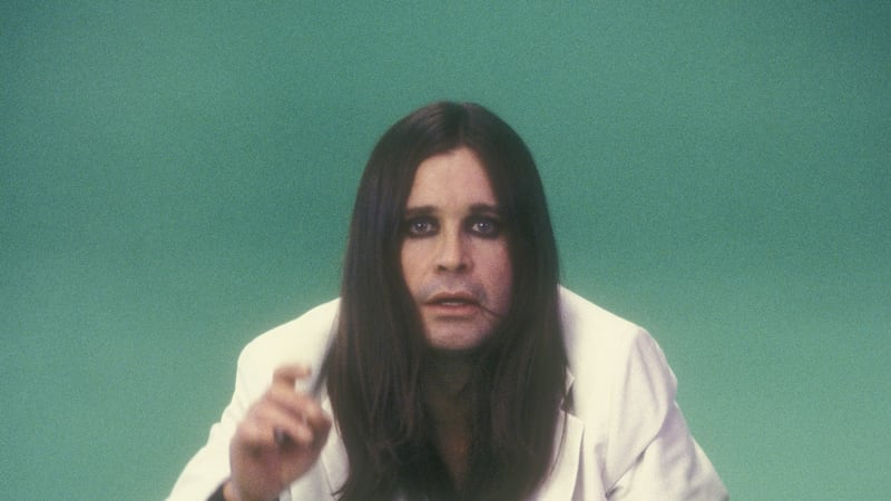 a vintage photo of rock star Ozzy Osbourne in a white suit with long straight black hair and black eyeshadow