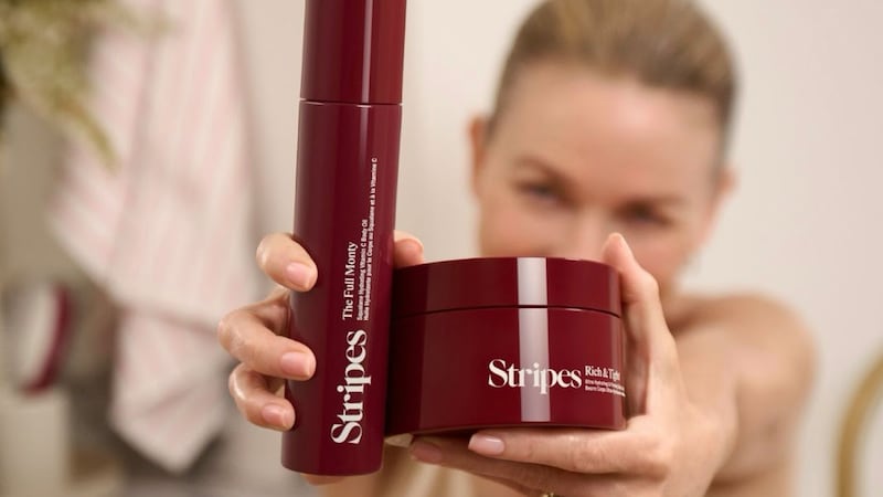 Stripes Beauty founder Naomi Watts is blurry in the background. in the foreground, her extended arm holds two Stripes Beauty products: a tub of lotion and a tall bottle of oil.