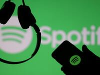 A smartphone and a headset are seen in front of a screen projection of Spotify logo.