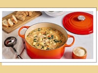Orange Le Creuset Dutch oven filled with creamy gnocchi soup, surrounded by sliced bread, grated cheese, and a ladle, styled on a kitchen table.