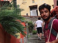 An image of Hersh Golberg-Polin, wearing an orange backpack, in front of a yellow building and stone steps.