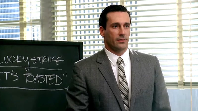 Don Draper (Jon Hamm) in Season 1 of "Mad Men" wearing a gray suit in front of a chalkboard that reads, "Lucky Strike, It's Toasted"