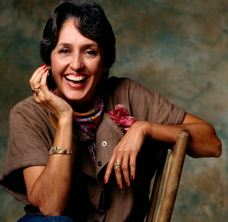 Musician Joan Baez sits sideways in a chair, with one arm on the back and the other holding her face, smiling at the camera in Los Angeles in 1983.