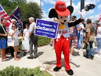 A person wearing a mouse costume holds a Governor Ron DeSantis poster