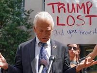 Peter Navarro, wearing a suit, stares down during a press conference where he was heckled by a protester holding a “TRUMP LOST” sign.