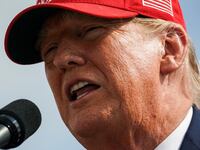 Donald Trump, wearing a red “Make America Great Again” hat, speaks on stage.