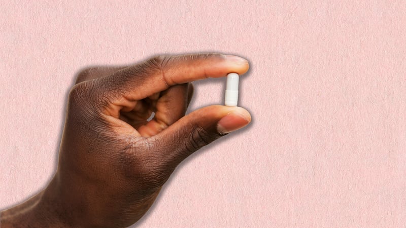 a hand holding up a small white supplement pill over a pink background