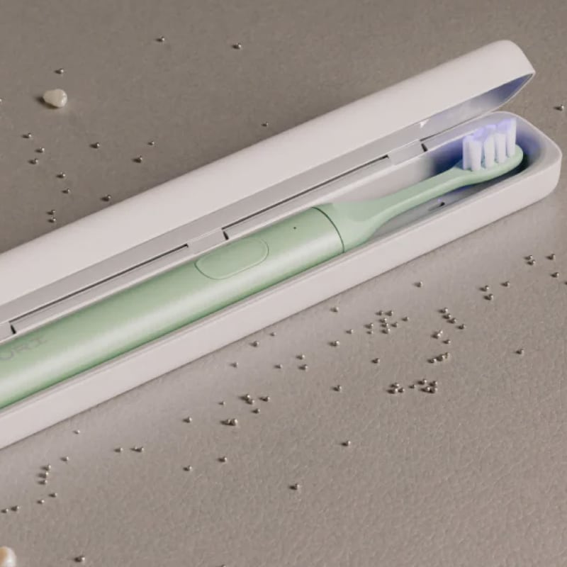 A mint green Suri electric toothbrush nestled inside an open white travel case, glowing with a soft blue UV light. The case rests on a grey surface scattered with small silver beads and shell fragments.