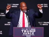 Mark Robinson raises his hands while standing behind a Trump-Vance branded podium.