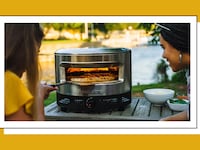 Two friends making a pizza outdoors with the Solo Stove Pi Prime pizza oven on a wooden table near a lake, stainless steel oven glowing with heat and fresh pizza baking inside.