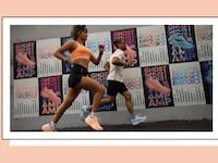 Two runners — a woman in a peach sports bra and black shorts, and a man in a white tee and black bike shorts, both in Brooks running shoes — jog past a wall covered in colorful Brooks "Ghost"/"AMP" ad posters, framed with a white and black border on a peach background.