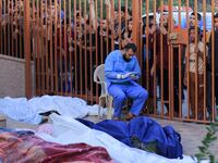 A medical worker sitting among bodies covered in white shrouds, with people looking over through a gate.