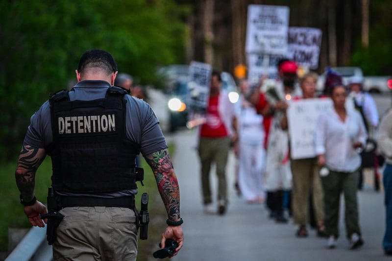 A corrections officer walks beside people holding candles, signs, and flowers during a vigil outside the Krome Detention Center in Miami on May 24, 2025, protesting US Immigration and Customs Enforcement custody and mass deportations.