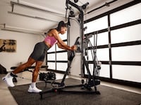 Woman performing a standing cable press on the BowFlex Xtreme 2 SE home gym in a garage gym setup, with a treadmill, dumbbells, and workout bench visible in the background.