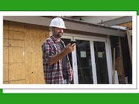 A construction worker wearing a white helmet looking at his phone. Behind him is a house under construction.