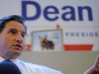Dean Phillips, wearing a shirt and tie, speaks during an interview, with a “Dean For President” sign in the background.