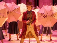 Timothée Chalamet as Willy Wonka stands in the middle of a pink room, in front of several people holding umbrellas that say “Wonka” on them.