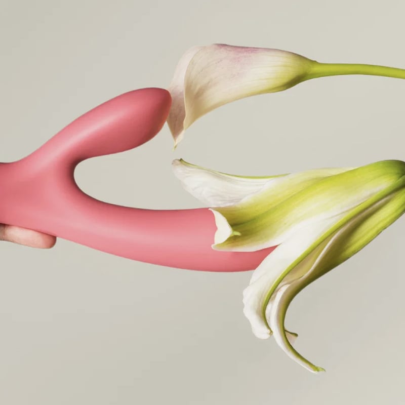 A hand grips a coral-pink rabbit vibrator as it meets a white calla lily, the two heads of the toy emerging from the flower's petals against a soft gray background.