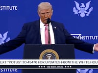 Donald Trump stands on stage at a lecture with his arms stretched out.
