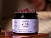 An open jar of CBDistillery Sound Sleep CBN gummies on a wooden nightstand beside a book, filled with sugar-coated black cherry gummies, with a softly blurred bedroom scene in the background