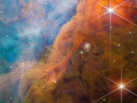 A nebula made of many layers of cloudy, colourful material. The top-left side of the image is brightly lit, filled with wispy, thin material in pale shades of pink and blue. A thick bar of denser, cloudier material crosses diagonally at the bottom right.