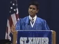 Vivek Ramaswamy standing at lectern behind St. Xavier sign, wearing academic robe and standing in front of American flag.