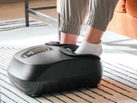 A person using the AuraHeal foot massager. They are sitting down on a couch and have both feet in the massager. They have grey pants and white socks on.