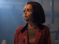 Emilia Clarke looks off into the distance, while wearing a red jacket in a dark room.