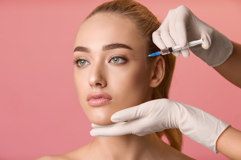 Young woman getting beauty injection near eyes, pink background