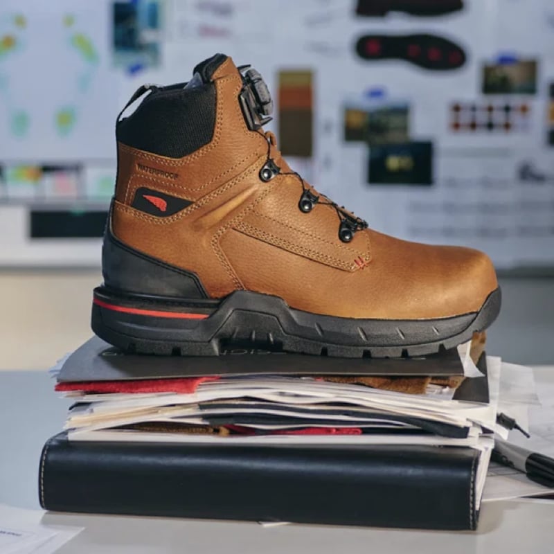 A Red Wing IronFlex work boot displayed on a stack of design documents in a footwear studio, with sketches, sole diagrams, and color swatches visible on a mood board in the background.