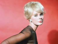 Elke Sommer, German actress, wearing a black dress with black chiffon sleeves in a studio portrait, against a red background, circa 1965.