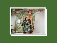 A woman wearing a dark navy satin cami romper and matching lace-trimmed robe covered in a leopard and green foliage print, standing in a room with chinoiserie-style wallpaper, a brass chandelier, and white paneled doors.