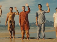 Five people stand on a beach while waving.
