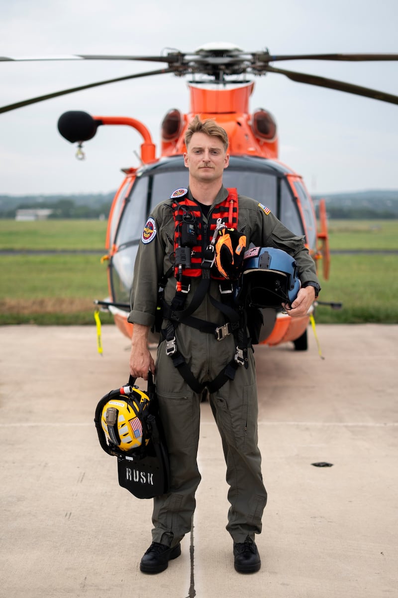 A man in flying uniform standing on tarmac in front of an orange helicopter. He is holding a flying helmet under his left arm and equipment in right hand.