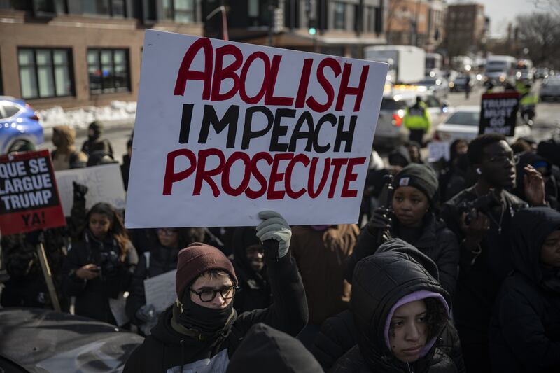 A masked man holds a sign in a crowd that says "ABOLISH, IMPEACH, PROSECUTE"