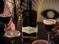 A table with four cocktails and a black bottle of Mr Black Cold Brew Coffee Liqueur on it. Two of the drinks are being held by hands.