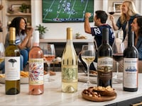 Five wines from Firstleaf sit on a counter with appetizers. In the background, friends cheer as they watch a football game.