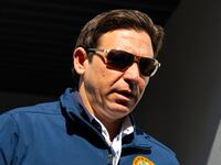 Ron DeSantis, wearing sunglasses, walks through a doorway while looking down.