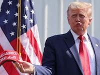 Donald Trump purses his lips while holding a red “Make America Great Again” hat on stage.